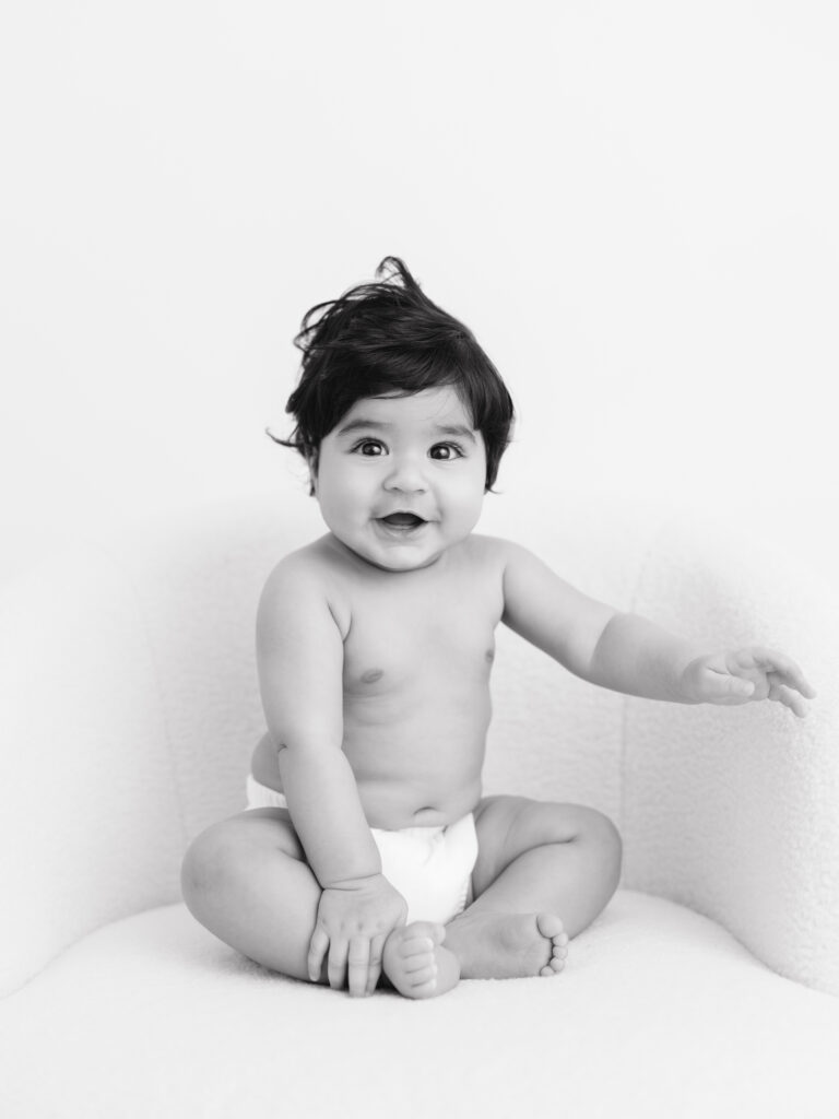 Baby sitting on a soft chair, smiling and reaching out, showcasing joyful expressions in a neutral-toned, minimalist setting, reflecting Sweet Jean Photography's focus on natural, timeless milestone sessions.