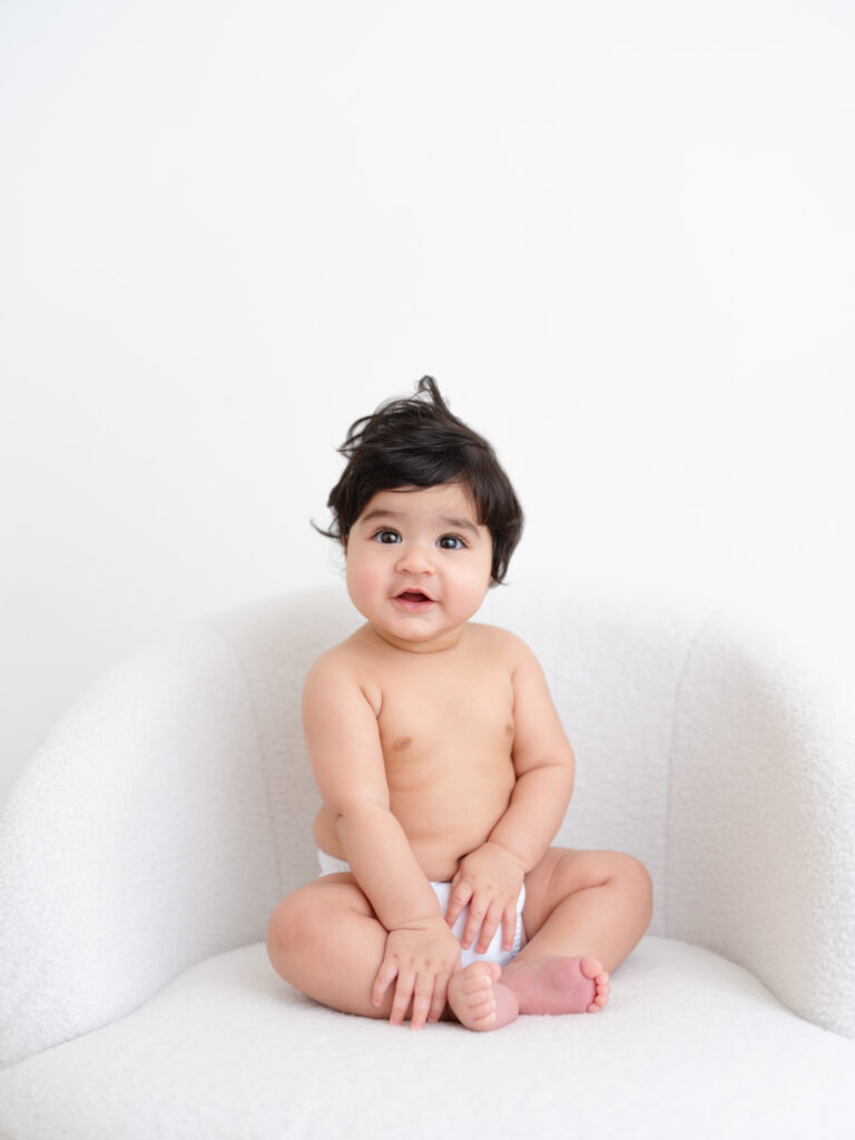 Baby sitting on a soft, neutral-toned chair, smiling with curly hair, capturing a joyful milestone moment in baby photography.