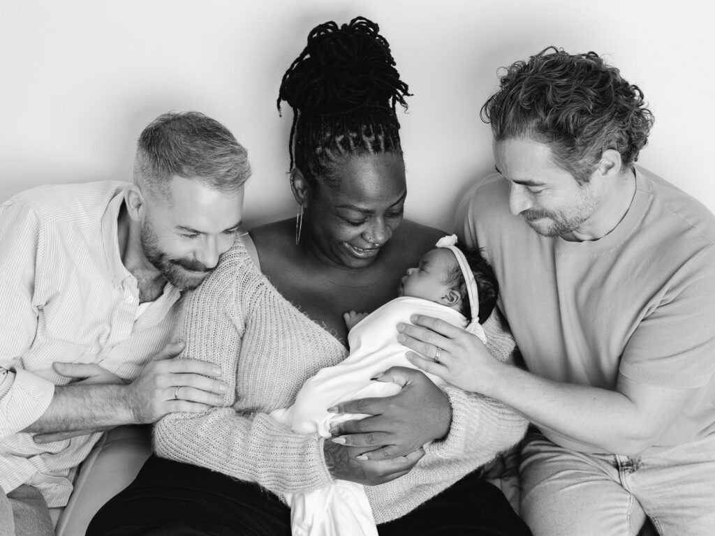 Two fathers and their surrogate holding a newborn baby during an intimate Sacramento family photography session in a light filled studio