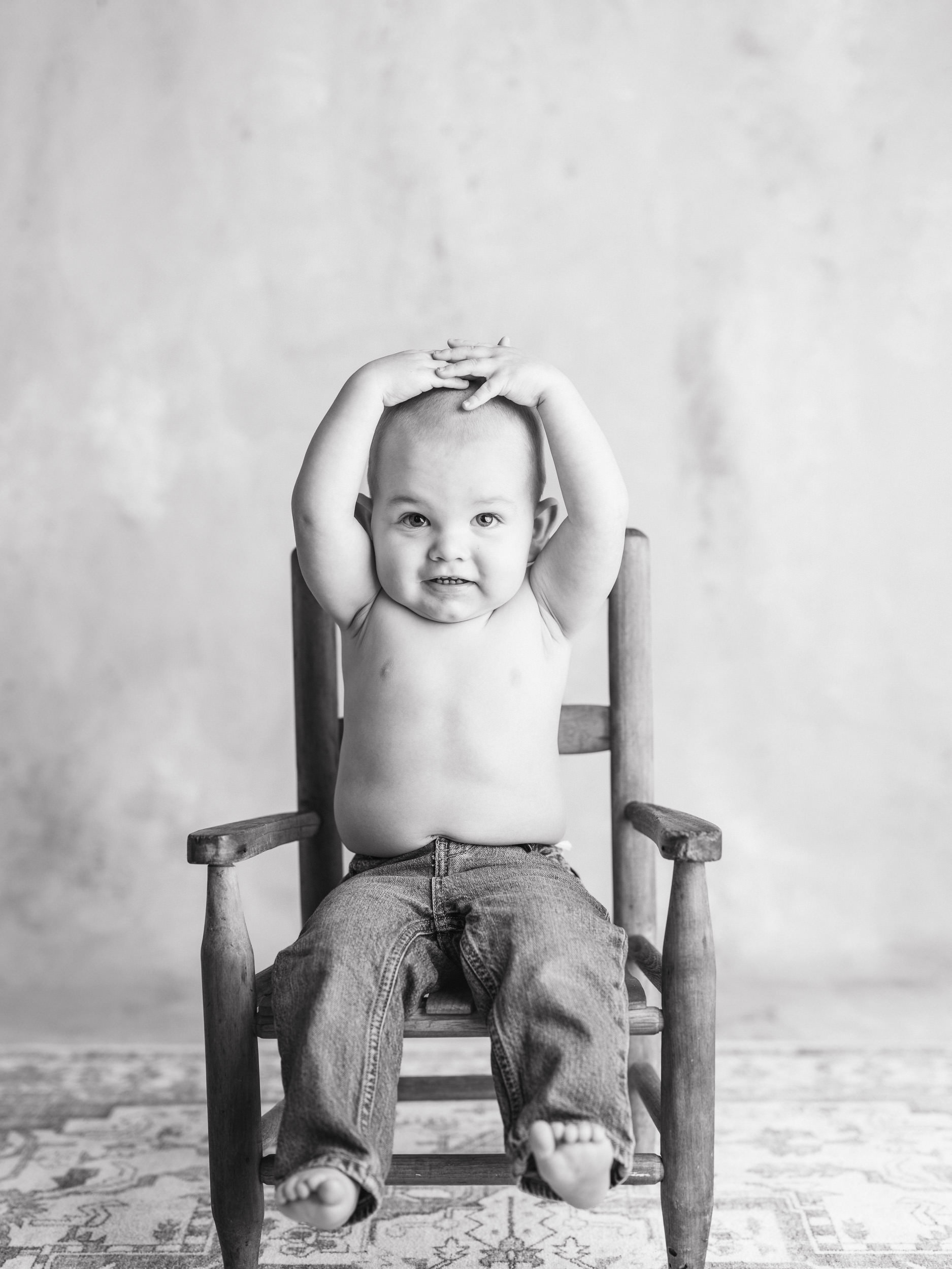 Fine art black and white studio portrait of a baby sitting in a wooden chair with arms raised, captured naturally for The Light Between childhood series.