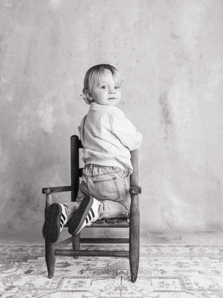 Fine Art Black and White Child Portraits Sacramento photographed in studio for The Light Between childhood series toddler sitting on a wooden chair, looking back over their shoulder.