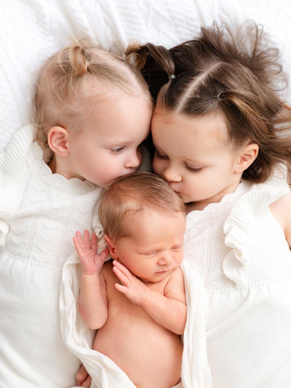 Sibling portrait of two sisters kissing newborn baby during Roseville studio session